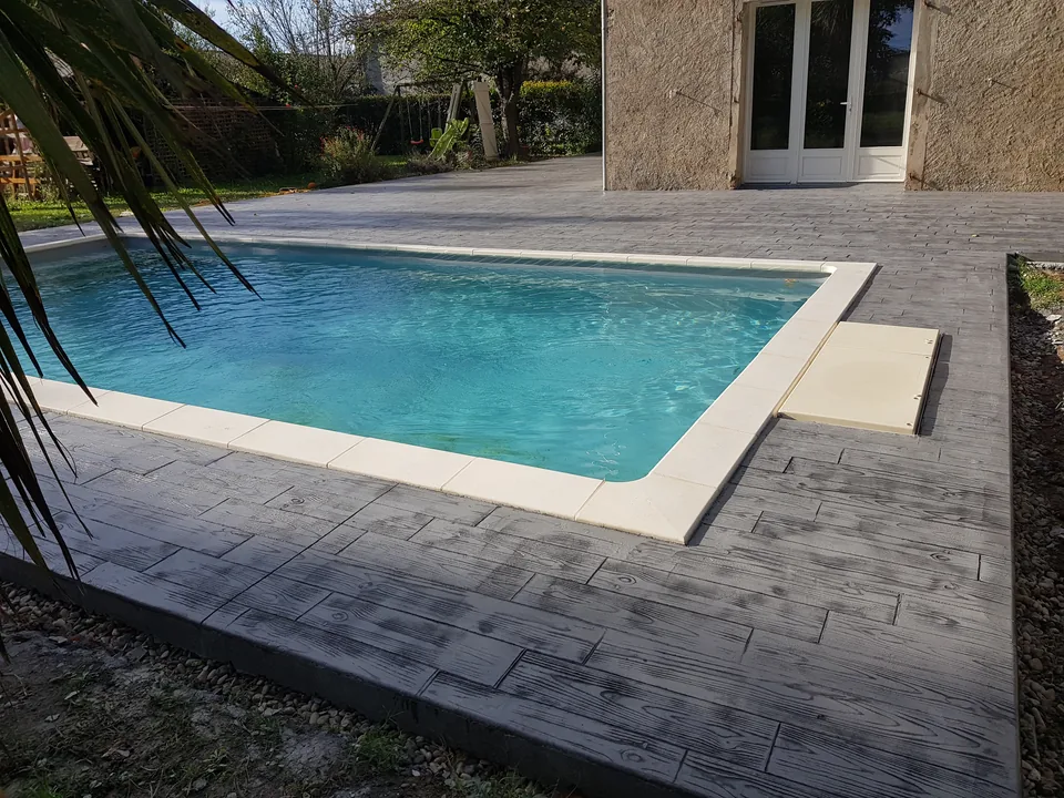 Béton imprimé gris à motif bois autour d’une piscine, devant une maison.